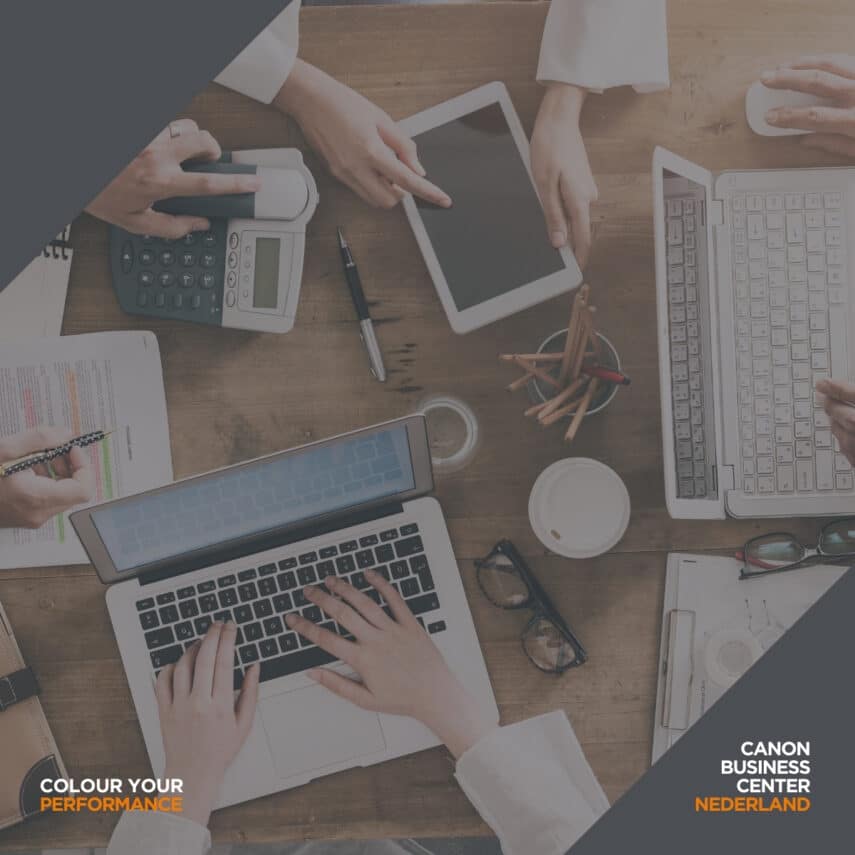 Bovenaanzicht van een houten bureau met laptops, een tablet, een rekenmachine, papieren en koffie, omringd door mensen die samenwerken.