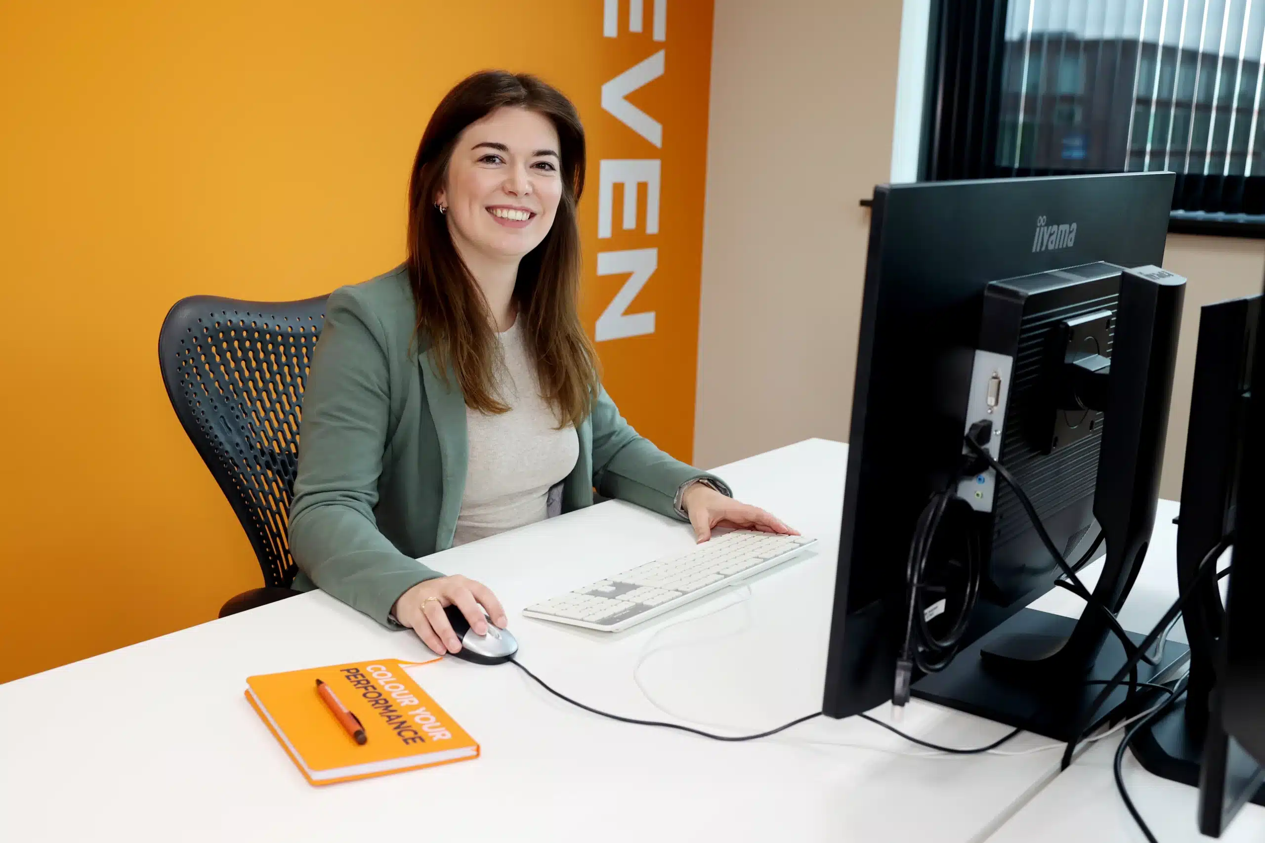 Marloes Brouwers aan het werk achter een computer, gekleed in een groene blazer, met een oranje boek op haar bureau.