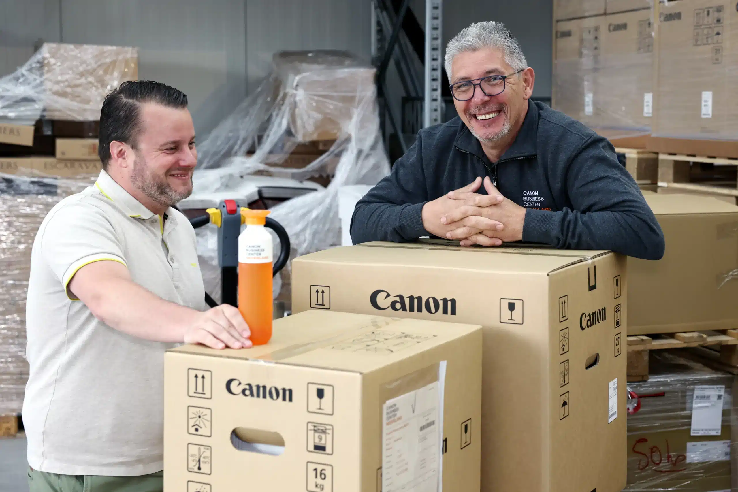 Twee collega's staan in een magazijn, leunend op grote Canon-dozen. De gezichten zijn geblurd. Op de voorgrond staat een oranje fles.