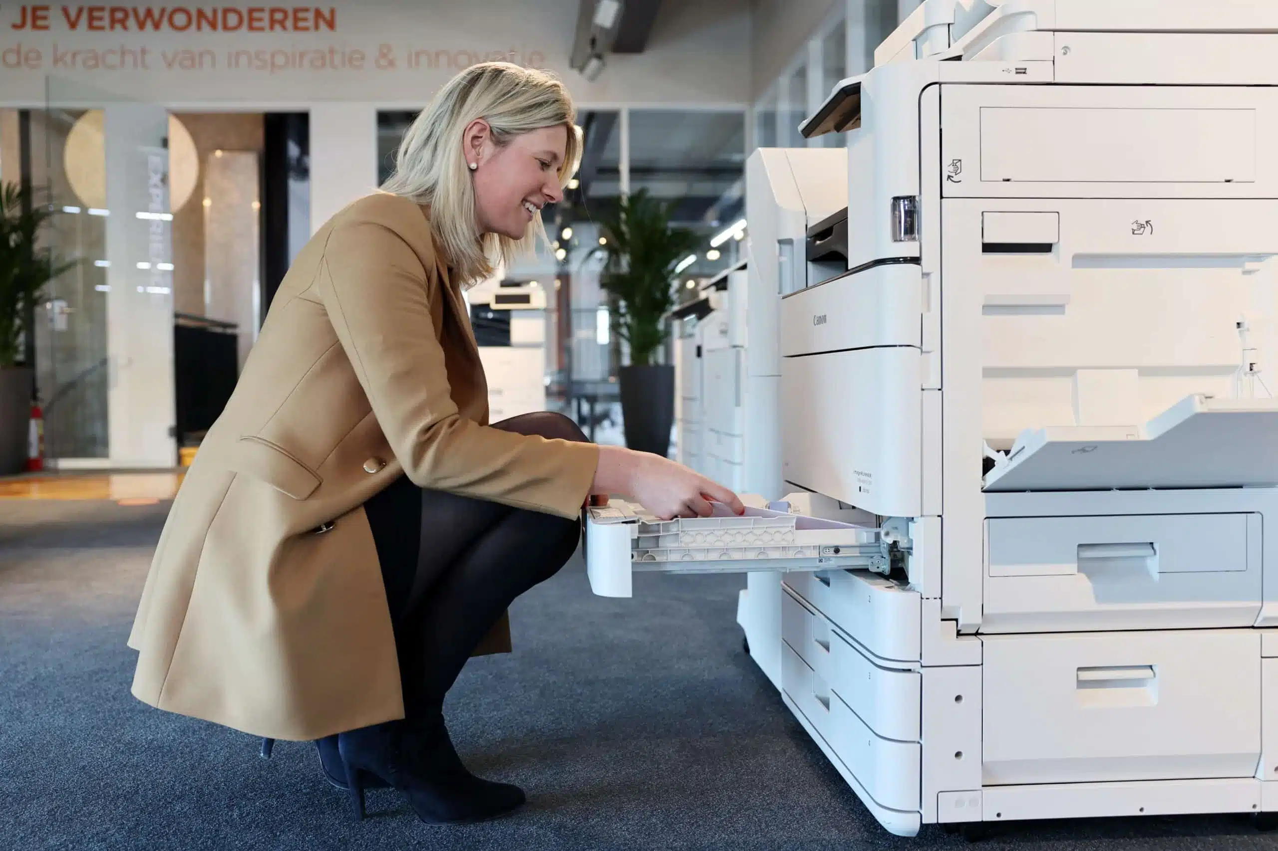 Een persoon in een bruine jas hurkt voor een grote witte printer en opent een lade — een visuele weergave van het gebruiksgemak van onze printoplossingen in een moderne kantooromgeving.