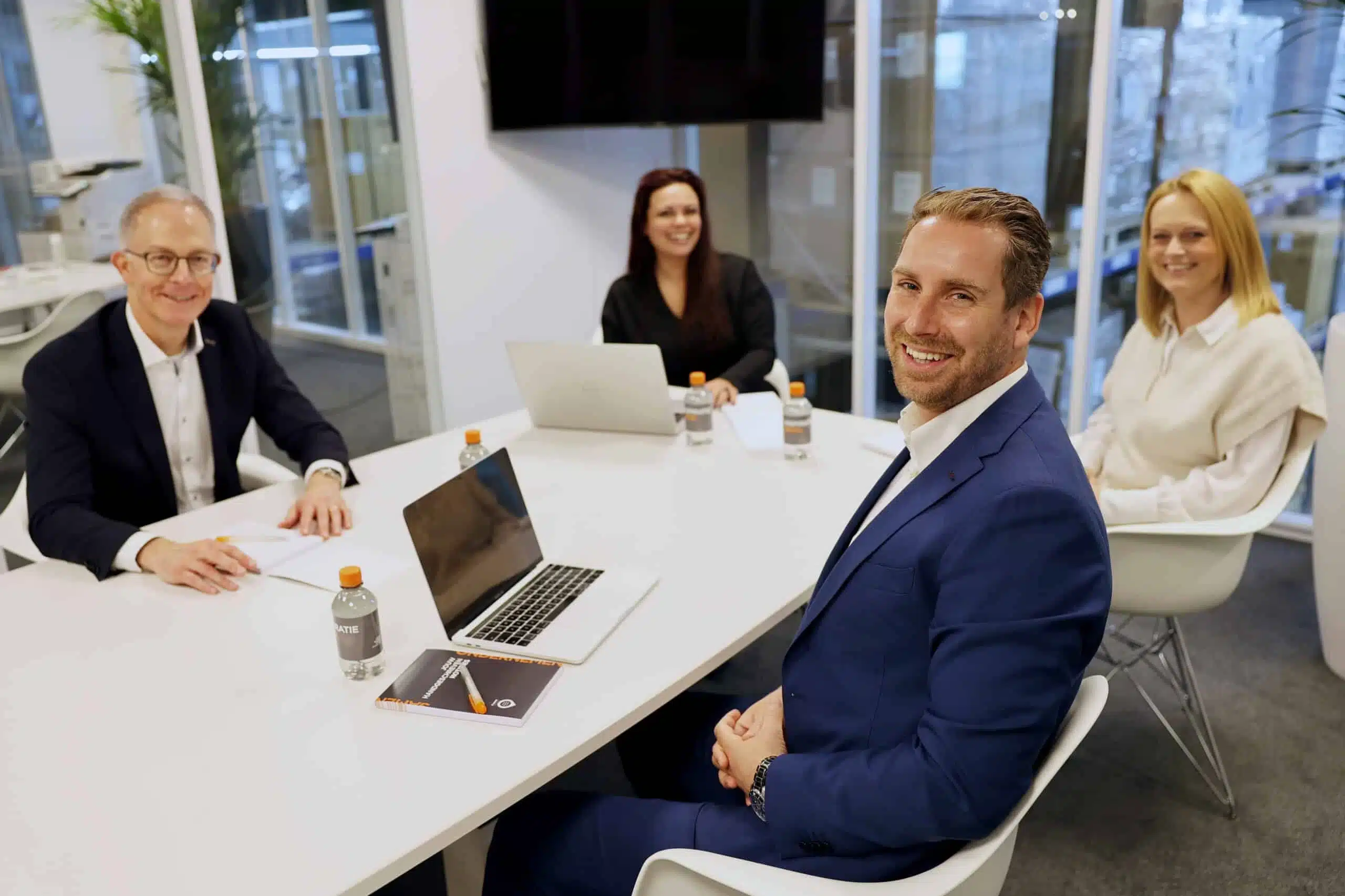 Vier mensen zitten rond een witte tafel in een moderne kantooromgeving met laptops en notitieboeken — een overlegsetting waarin onze printoplossingen onderdeel zijn van een bredere digitale werkomgeving.