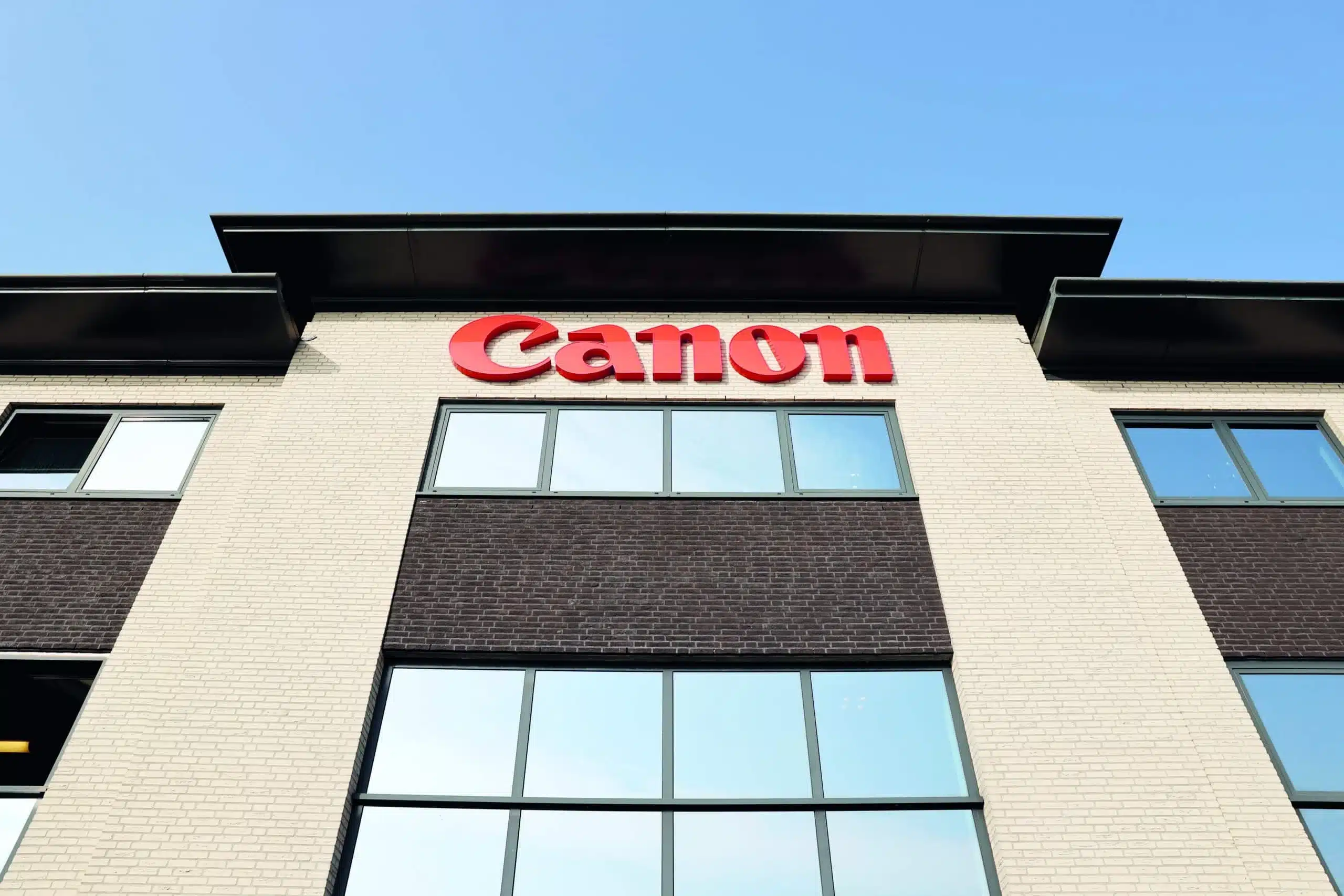exterieur van een gebouw met het canon-logo bovenaan, beige bakstenen gevel, grote ramen en een sectie met donkere stenen, onder een heldere blauwe lucht.
