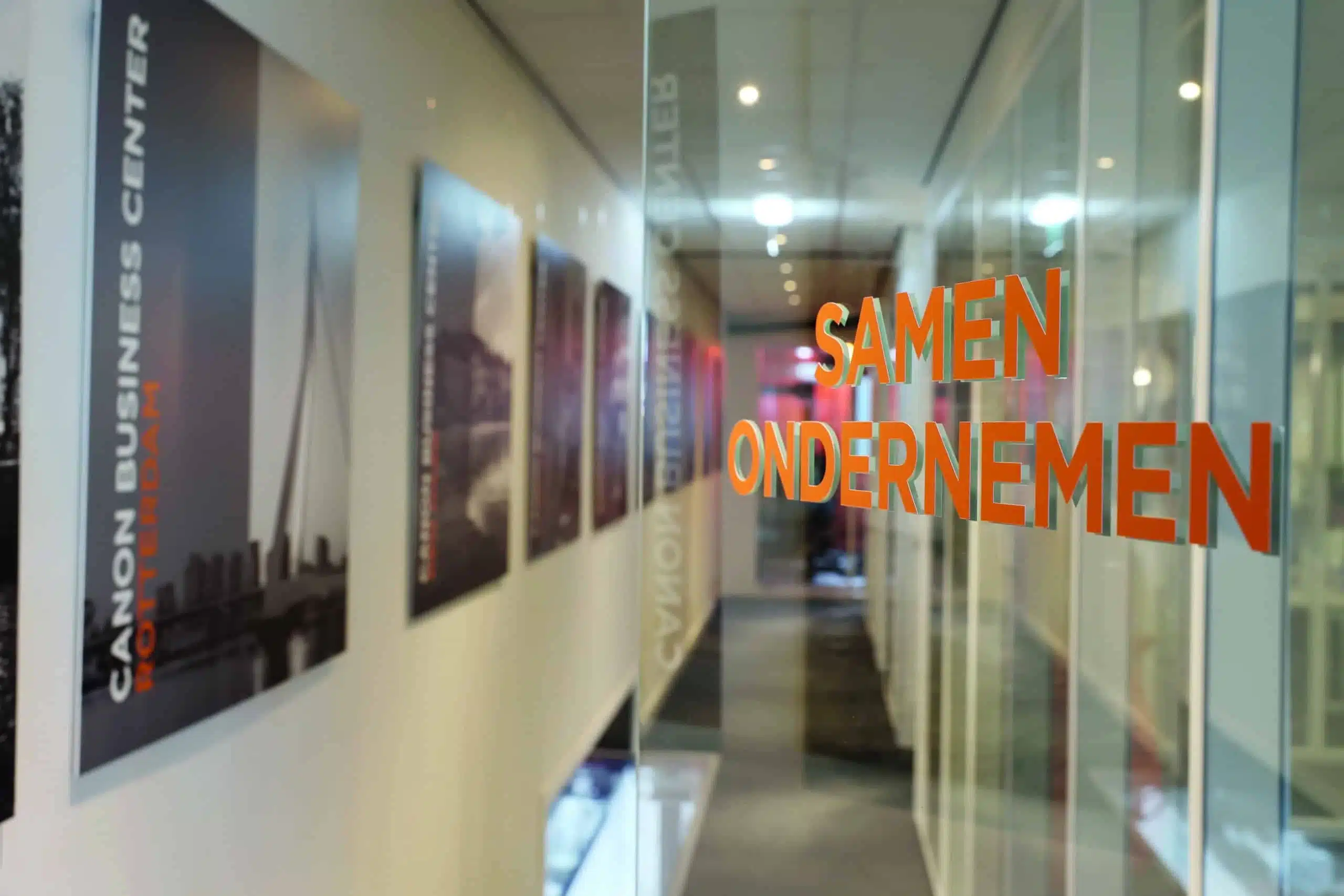 een gang met glazen wanden en ingelijste foto's, waaronder één met de tekst "over canon business center rotterdam", en de slogan "samen ondernemen" in oranje op het glas.