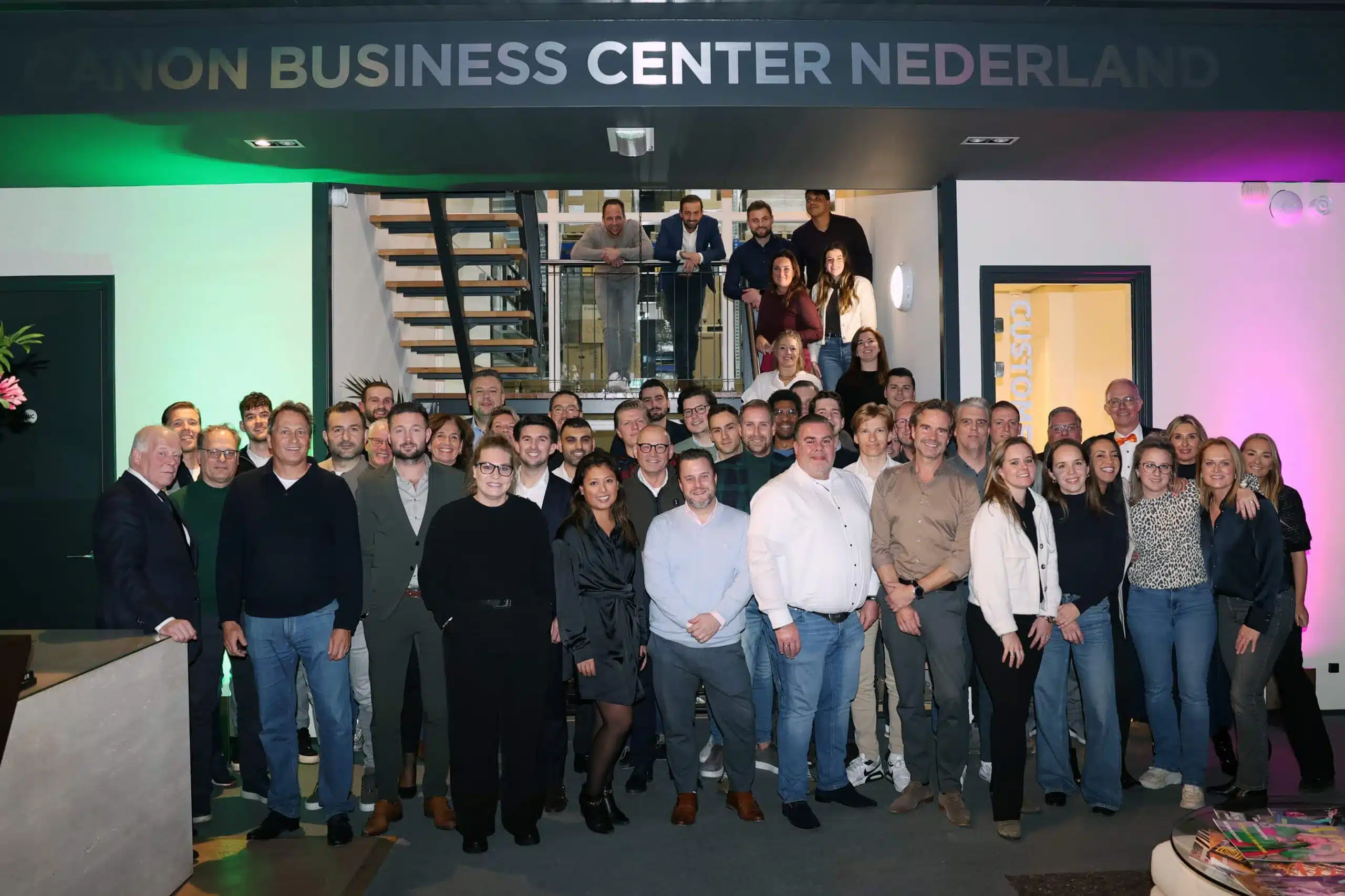Een groep mensen poseert voor een bord met de tekst "CANON BUSINESS CENTER NEDERLAND" in een kantooromgeving met een trap en mensen op de achtergrond.