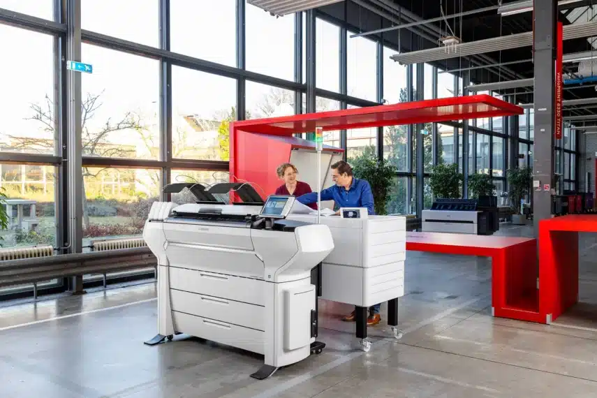 Twee mensen in een moderne kantoorruimte naast een multifunctionele professionele printers, omgeven door een industriële inrichting en kantoorapparatuur.