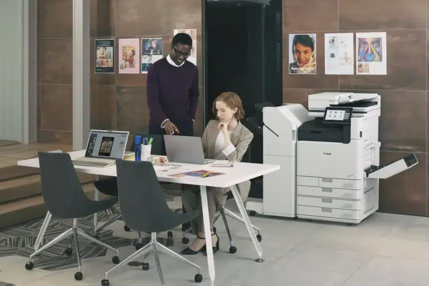 Twee mensen in een kantooromgeving, werkend aan een tafel met laptops en documenten, naast professionele printers.