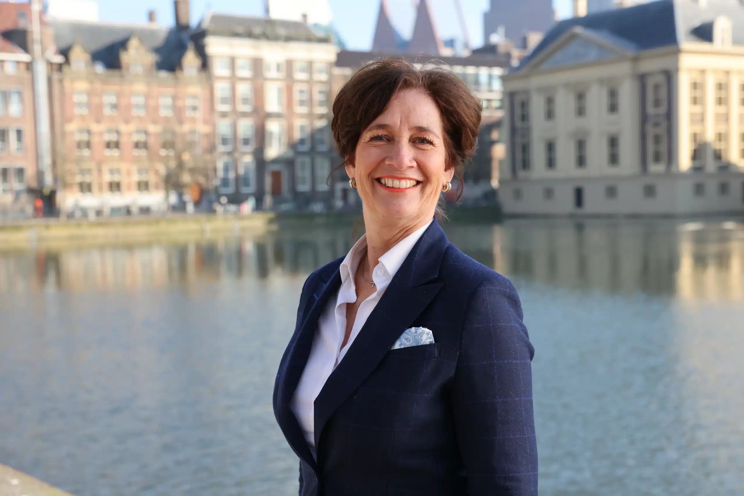 Sandra Vos in zakelijk kostuum lachend voor het Binnenhof in Den Haag, Nederland, op een zonnige dag.