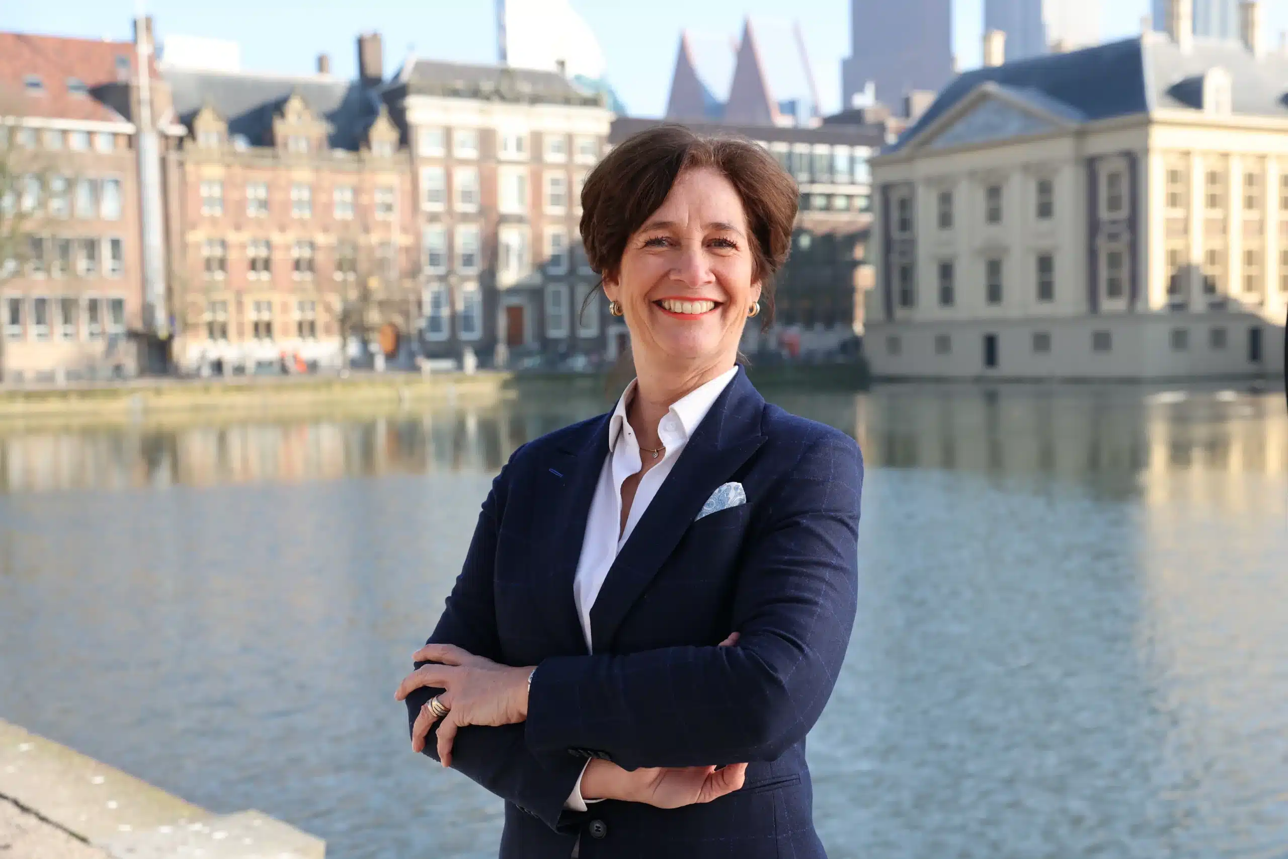 Sandra Vos kijkt glimlachend met armen over elkaar voor het Binnenhof in Den Haag met moderne skyline op de achtergrond.