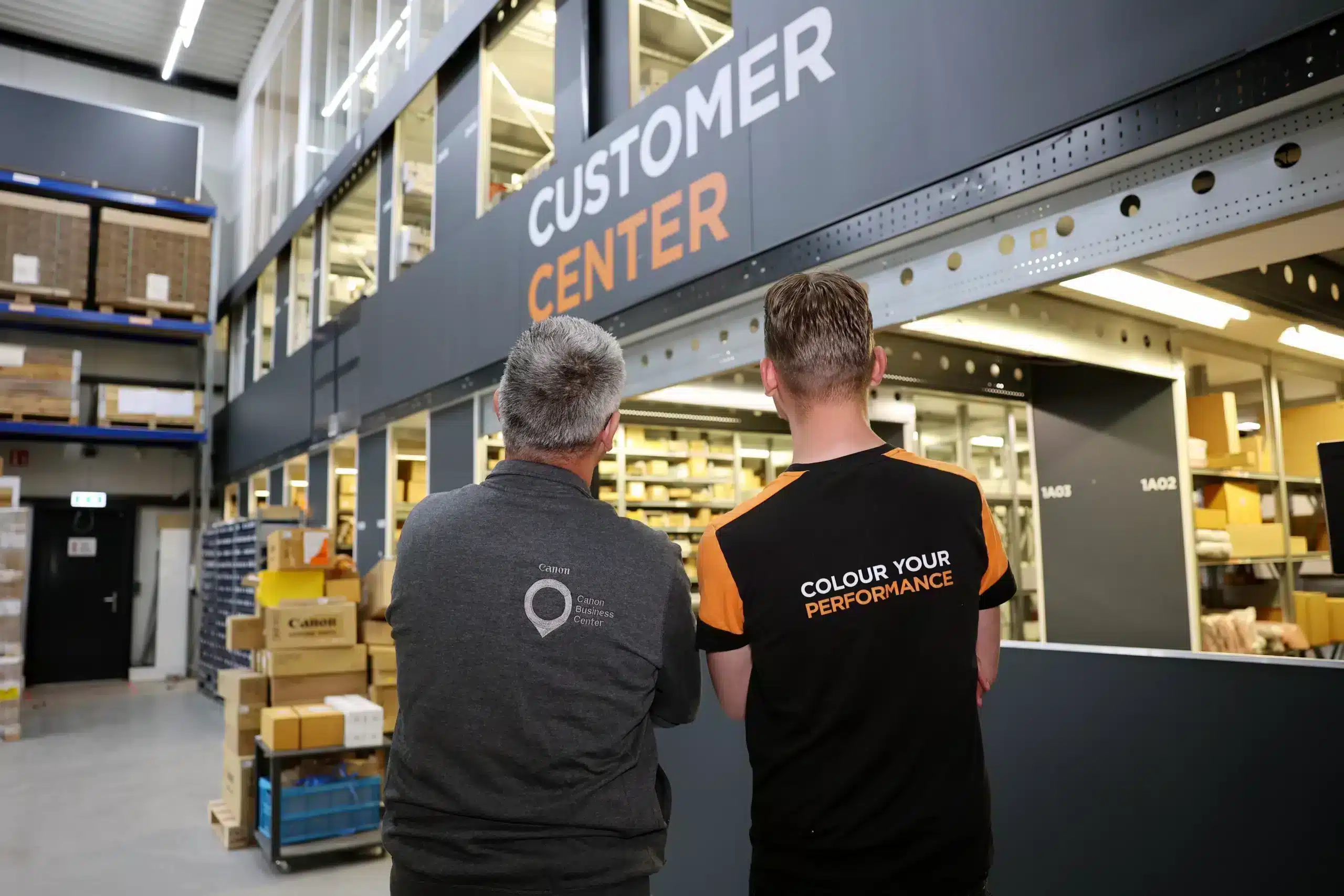 Offerte aanvragen: twee personen bij het CUSTOMER CENTER in een magazijnomgeving met dozen en stellingen.