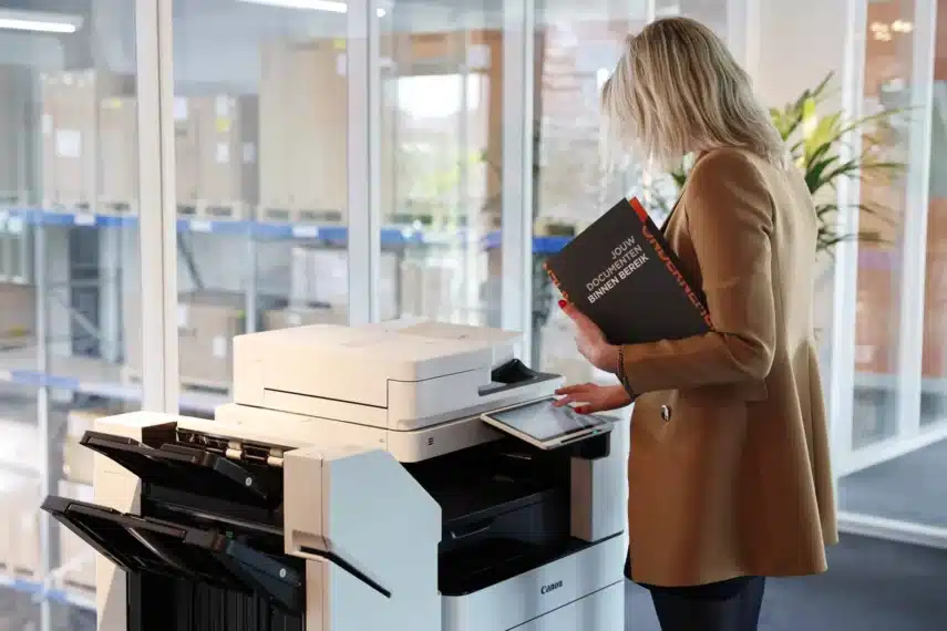 Vrouw bedient Canon grootformaat printers in een moderne kantooromgeving. Ze houdt een geprint document vast met de titel "Jouw Document Binnen Bereik" en navigeert via het touchscreen. Op de achtergrond zijn glazen wanden en opslagrekken zichtbaar.