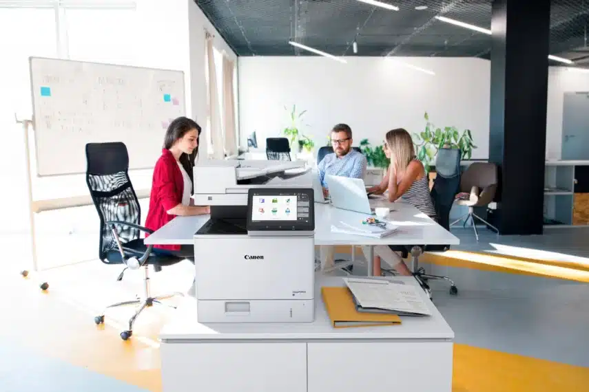 Nieuwe printer kopen voor kantoor? Moderne Canon printer op een bureau in een open werkruimte met medewerkers en laptops.